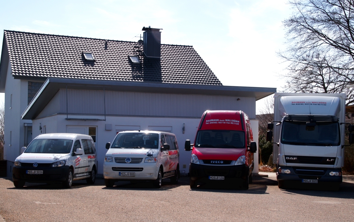 vier-Fahrzeuge-der-Firma-Baumann-Kurierdienste-stehen-in-einer-Reihe-vor-weissem-Gebaeude